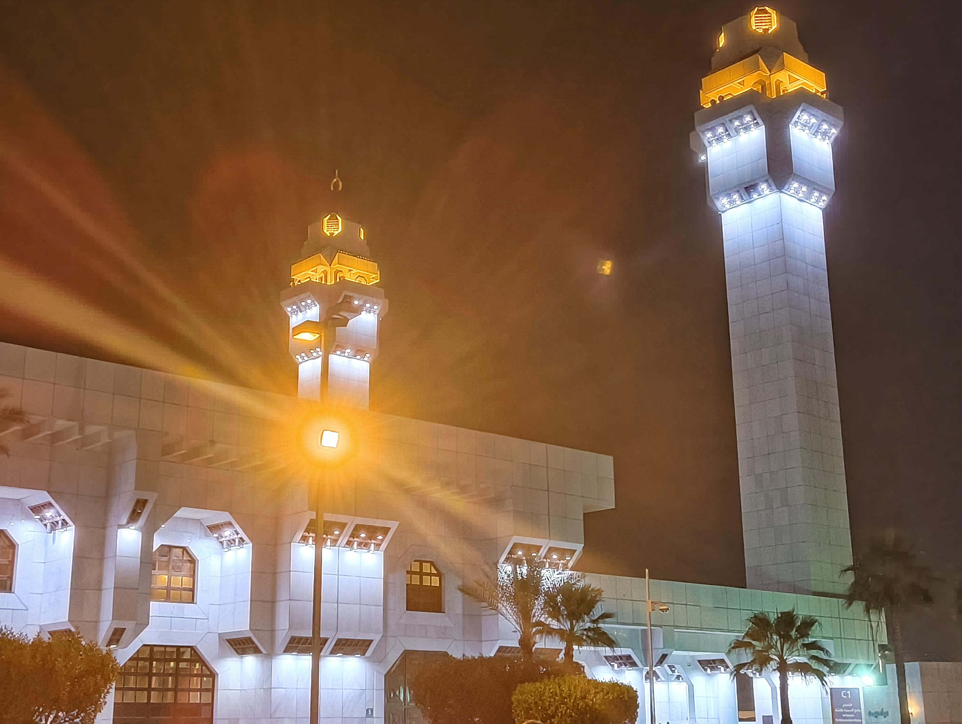 A beautiful shot of Masjid-e-Ayesha (Ayesha's Mosque) In Mecca, Saudi Arabia. lit up with blue lights at night, surrounded by palm trees. The foreground has a park area with warm-toned lighting from street lamps, creating a lovely contrast.