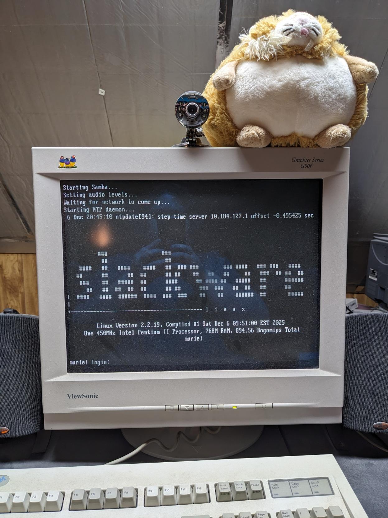 A beige CRT monitor displaying a near-fullscreen ASCII art rendition of the Slackware Linux logo, a brief summary of system info, and the login prompt. There is a silver webcam and a sabertooth cat plushie sitting atop the monitor.