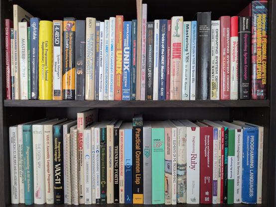 Two shelves of books.  The top shelf is books about operating systems, with CP/M, BSD, Unix, VMS, and Xinu represented, followed by some texbooks on operating systems and a somewhat out of place programming encyclopedia.  The lower shelf is programming language books, starting with various assembly languages (PDP-11 and VAX), then Algol 68, Applesoft, AWK, BASIC, C, C++, Forth, Icon, Lisp (lots of Lisp), ML, Prolog, Ruby, Scheme, POSIX shell, Smalltalk, Squeak, and a couple of books covering a variety of languages.  Again, a somewhat out of place copy of Kernighan and Plauger's Software Tools closes out the shelf.  Most of the books are late 1980s or older, although some thoroughly modern books such as Seibel's Practical Common Lisp and Hoyt's Let over Lambda do make an appearance.  Nestled in in the middle of the top shelf are the edges of an original copy of the Lions Commentary, both volumes in UNSW covers, immediately beside a modern commercial reprint.  You can't see it from here, but both the hardback version of The Unix Programming Environment and the second edition of The C Programming Language are signed by Brian Kernighan.