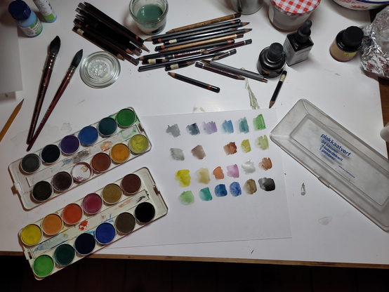 Photo of a drawing desk. In the middle is a piece of Bristol board with color swatches. Left are two trays of twelve cups a tray of gouache paint. Right is the lid of the painting box, with the text "Plakkaatverf. 24 colors.1 tube of covering white.1 brush". The cover is transparent for the rest.

North of the paper with color swatches are a mess of pencils, west are two big covering French (but US-made...) aquarel brushes, east are bottles of India ink.