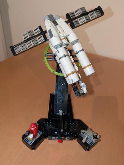 Fully assembled Lego model on a brown table. It consists of a black tilted base that holds a mostly white spaceship. The ship has three massive tanks / engines, and two massive solar panels to the sides.
The base has two minis standing on it. A human in a red space suit on the left, and a brown, round, five armed / legged creature on the right.