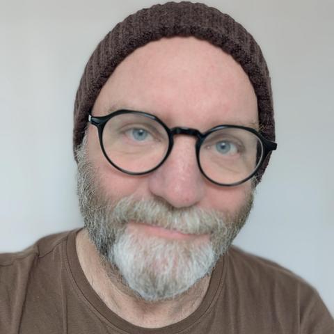 Photographic portrait of a middle-aged man with a grey beard, spectacles, and a woolly beanie hat.