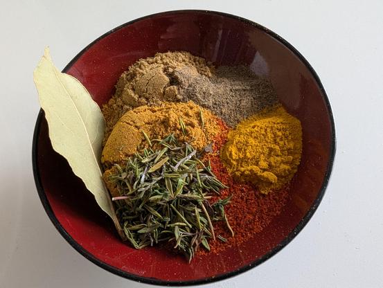 A small red and black bowl filled with a pile of herbs and spices in different colours (a bay leaf, dried rhyme, cumin, sweet mix curry powder, smoked paprika, ground black pepper and turmeric)
