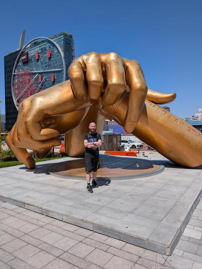 I am standing in front of a sculpture of two hands, crossed and doing the horse riding pose from the song Gangnam style. I'm looking at the camera and trying to do the same pose. Trying.