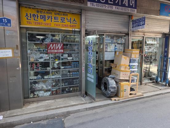A tow of small shops. The first one has Mean Well power supplies on display. The second one has a lot of boxes in front of it and it is not clear what it sells. And the third one sells valves I think.