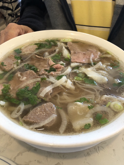 close-up view of a large white bowl filled with noodle soup. The soup contains white rice noodles, slices of cooked meat, sliced white onions, and chopped green herbs in a clear, brownish broth. A hand is partially visible on the left side of the bowl. The bowl rests on a light-colored, marbled surface. In the background, a person is wearing a dark jacket and a yellow and white striped shirt.