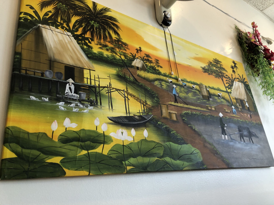 landscape painting depicts a rural scene under a bright yellow and orange sky. In the foreground, large green lily pads with white flowers float on the water. On the left, a wooden hut on stilts sits above the water near a person wearing a conical hat and several white birds. A small dark boat floats in the middle ground. To the right, a path leads past small huts toward a person plowing a field with a large, dark animal.