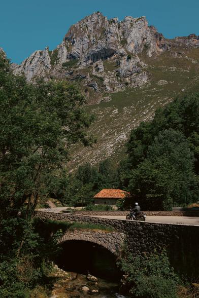 A vertical photo of a motorcyclist riding over a bridge made of stones with a stone house with a red rood behind and tall rocky mountains towering above. Green leafy trees on either side of the photo.