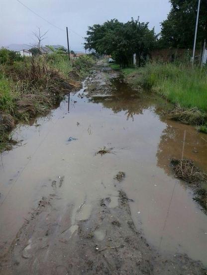 image shows a severely damaged, unpaved rural road flooded with large, stagnant puddles of brown water.