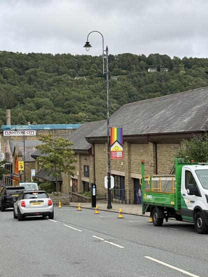 A street view with cars and traffic cones. In a tall lamppost is the Pride flag with the words “Report Hate Crime” and a QR code on the bottom