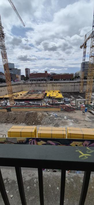 An elevated view overlooking an active urban construction site framed by two large yellow cranes on the left and right edges. In the foreground, a dark railing with yellow graffiti sits above stacks of bright yellow rectangular blocks and piles of dirt. The center of the image shows a deep excavation area containing scaffolding, wooden forms, and various metal structures behind a green fence. A cityscape with several buildings and a bridge is visible in the background under a cloudy sky. The two tall yellow cranes extend upwards toward the top of the frame.