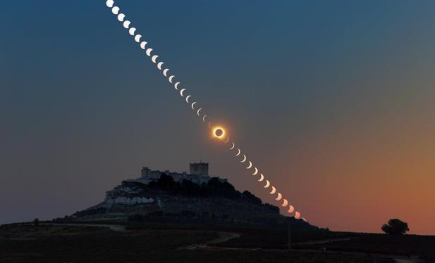 A photograph of a dimly lit castle silhouetted against the dusk sky. It's a combination of multiple exposures, with the Sun appearing as a sequence of discs crossing the sky diagonally above the castle. A progressively smaller fraction of the Sun is visible as the eclipse progresses. Right above the castle the eclipse reaches the total phase, with the Sun completely blocked by the Moon, revealing a fiery corona around it. The sequence continues towards the horizon, with the Sun progressively reappearing.