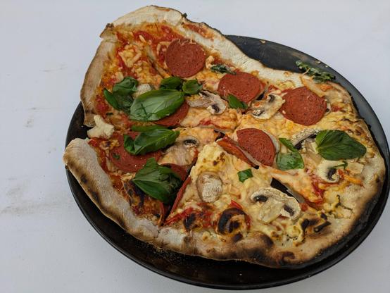 A freshly cooked homemade pizza on a black plate, topped with mushrooms, cheese, basil and round slices of topping, with a charred and crisp pizza crust.