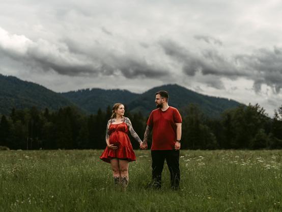 An outdoor portrait in a lush green meadow in Bavaria, captured by European elopement photographer Sturmsucht, featuring a pregnant woman and a man holding hands while looking affectionately at each other. The woman, on the left, has blonde hair tied back and is wearing a rust-red spaghetti-strap maternity dress with visible tattoos on her arms and legs, her left hand resting gently on her baby bump. The man, on the right, wears glasses, a dark red t-shirt, and dark trousers, with tattoos covering his arms. They stand amidst tall grass and wildflowers under a dramatic, overcast sky filled with heavy grey storm clouds, backed by rolling green hills and a dense dark forest.