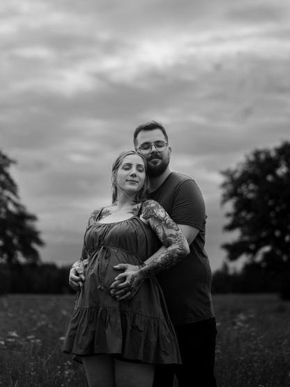 An intimate, black-and-white portrait captured in Bavaria by European elopement photographer Sturmsucht, showing an expectant couple standing in a grassy field with their eyes closed in serene contentment. The bearded man with glasses stands directly behind the pregnant woman, wrapping his arms securely around her waist as they both place their hands tenderly over her rounded baby bump. The woman wears a ruffled strap dress, her shoulders and arms heavily adorned with detailed tattoos that contrast softly against the muted tones of the open landscape and the expansive, cloudy sky above.