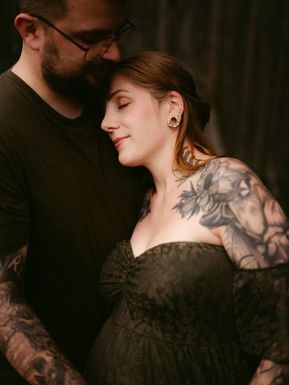 A close-up portrait in Bavaria by European elopement photographer Sturmsucht, capturing a tender moment between an expectant couple with their eyes closed in quiet connection. The pregnant woman rests her head gently against the man's chest, wearing an off-the-shoulder dark olive-green dress with intricate floral and botanical tattoos covering her arms and shoulder, complemented by ornate golden earrings. The bearded man, wearing glasses and a dark t-shirt with tattooed arms, leans in closely, resting his head softly against hers in an atmosphere of warmth and intimacy.