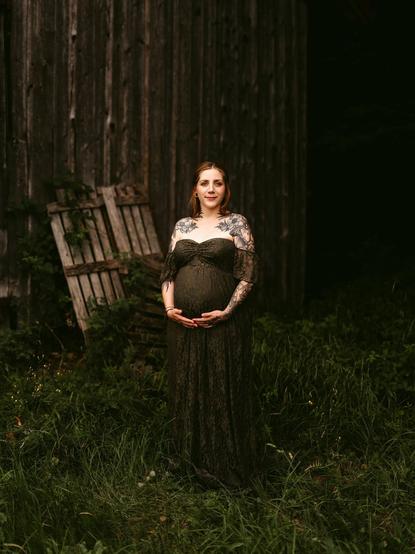 A full-length portrait in Bavaria by European elopement photographer Sturmsucht, featuring a pregnant woman standing gracefully in tall grass and greenery in front of a rustic wooden barn. She wears an off-the-shoulder dark olive-green lace maternity dress, gently cradling her rounded baby bump with both hands as she looks forward with a soft, serene expression, her shoulders and arms adorned with detailed floral and botanical tattoos.