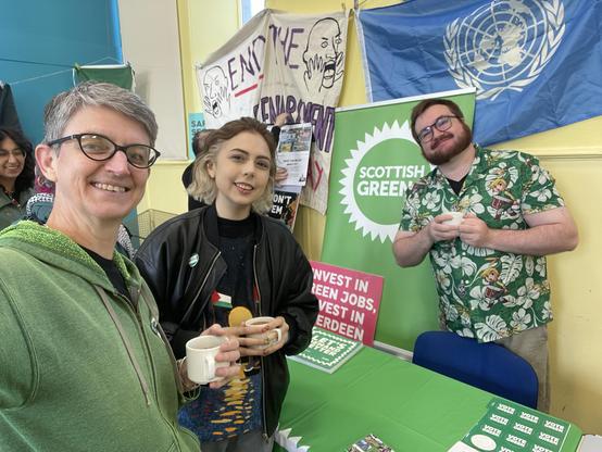 Maggie wearing a green hoodie with two other greens, at the Scottish Greens stall