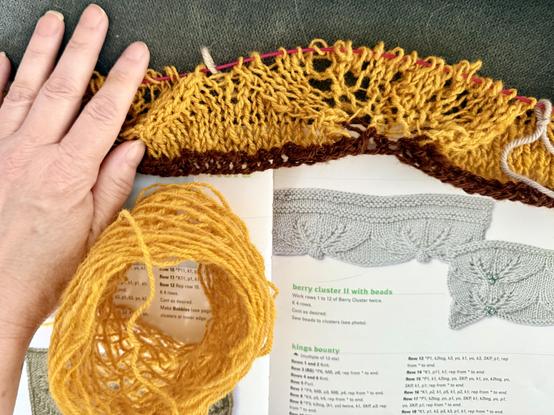 My hand holding a piece of yellow lace with a brown stripe on the edge. It's on a pink cable needle still and is resting on an open stitch pattern book that shows the lace pattern two ways. The lace is supposed to look like a cluster of three berries with either a bobble or beads where they meet in the center of the indented part of a scallop. Also shown is the remains of the yellow cake of yarn that's so depleted it looks lacy also.