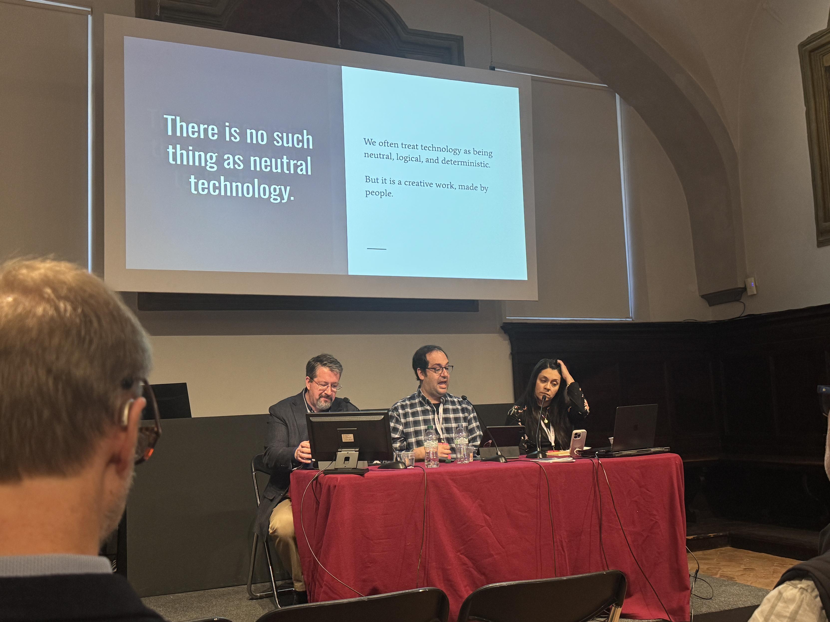 Two men and a woman on stage at a table in front of a slide with the text "There is no such thing as neutral technology.

We often treat technology as being neutral, logical, and deterministic.

But it is a creative work, made by
people."