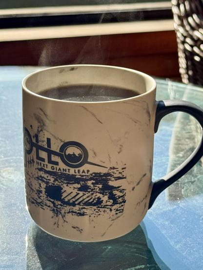 A large coffee mug with NASA Apollo Mission graphics on the side. On a glass table, steaming in the morning sun.