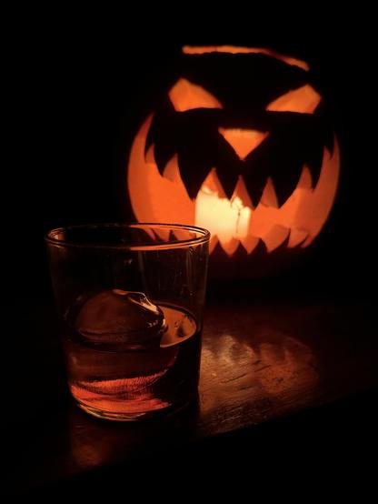 A short glass of whiskey in front of a glowing jack-o-lantern.