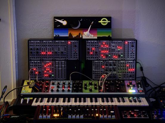 A Moog Matriarch synthesizer (and 5 octave keyboard) with a Roland System 100 clone from Behringer stacked on top, with sliders glowing red. On top of this is a lego set "Tales of the Space Age" with various space scenes. 