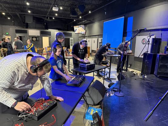 A room with a dozen people, some in the foreground with headphones, using some electronic musical instruments. 