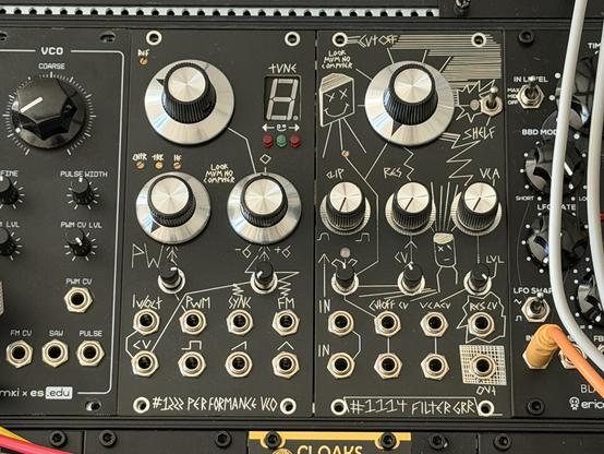 Close-up of a modular synthesizer rack with black panels covered in hand-drawn white graphics and labels. Large silver knobs dominate the center modules, surrounded by smaller black knobs, toggle switches, patch points, and a small digital numeric display. 