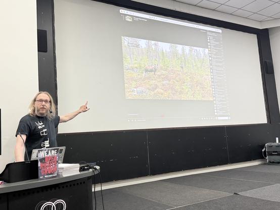 Michael Dales pointing at a slide showing moose captured in a forest on a TV camera.