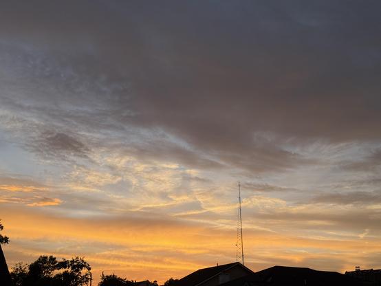 A cloudy sky at sunset. In the distance, a dark horizon with some trees in buildings and a tall radio tower.