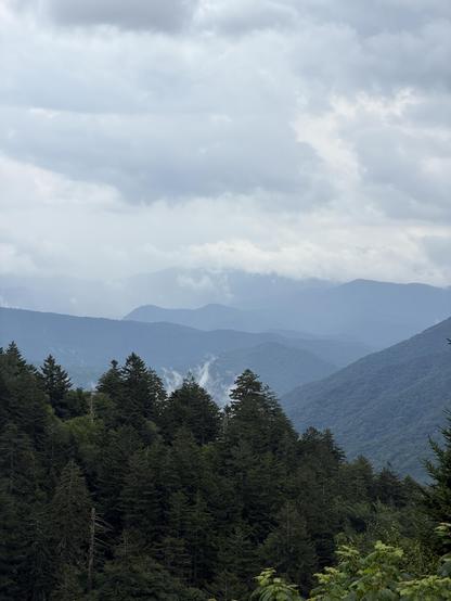 A picturesque view of the great Smoky Mountains national Park, near newfound gap. In the fore ground, lush green trees, and in the background, pale blue mountains, surrounded in white clouds.