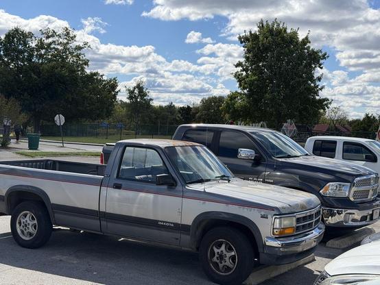 Two Dodge trucks, an 80's Dakota parked next to a modern RAM 1500. The Dakota is old but in decent shape. It's dwarfed by the new 'small' truck.